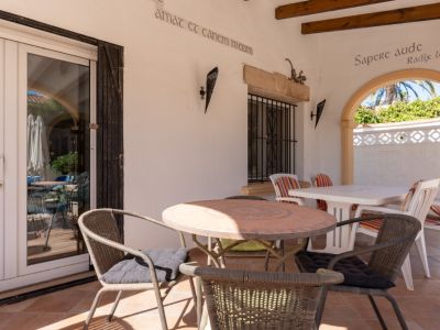 Terraza cubierta con suelos de terracota, mesa redonda de mosaico y puertas de PVC blanco. Un arco revela vegetación exterior y palmeras.