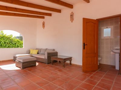 Terraza cubierta con suelos de terracota, vigas de madera y un sofá de ratán. Incluye un acceso a un aseo y una ventana arqueada con vistas a la vegetación.