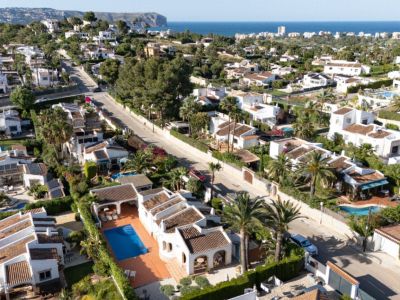 Vista aérea de la tranquila zona residencial de Jávea, mostrando villas blancas con piscinas privadas, exuberantes jardines mediterráneos y el mar azul a lo lejos.