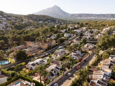Vista aérea de una tranquila zona residencial, mostrando numerosas villas blancas con tejados de terracota, enclavadas entre abundante vegetación mediterránea y calles serpenteantes, con una montaña destacada al fondo.