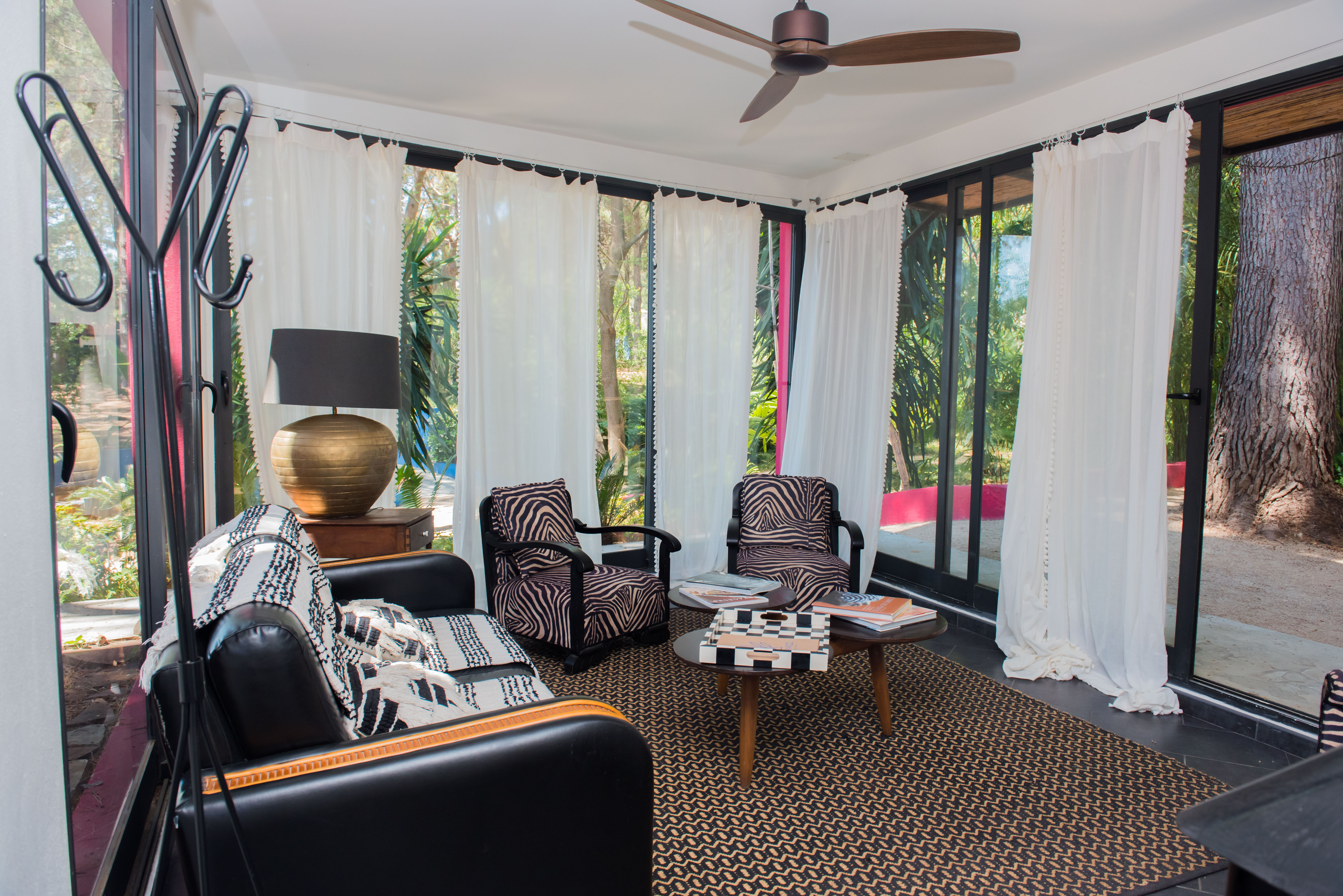 Acogedora sala de estar con amplios ventanales que enmarcan vistas a un jardín tropical. Incluye sofá de cuero negro, sillones con estampado de cebra, alfombra geométrica y ventilador de techo.