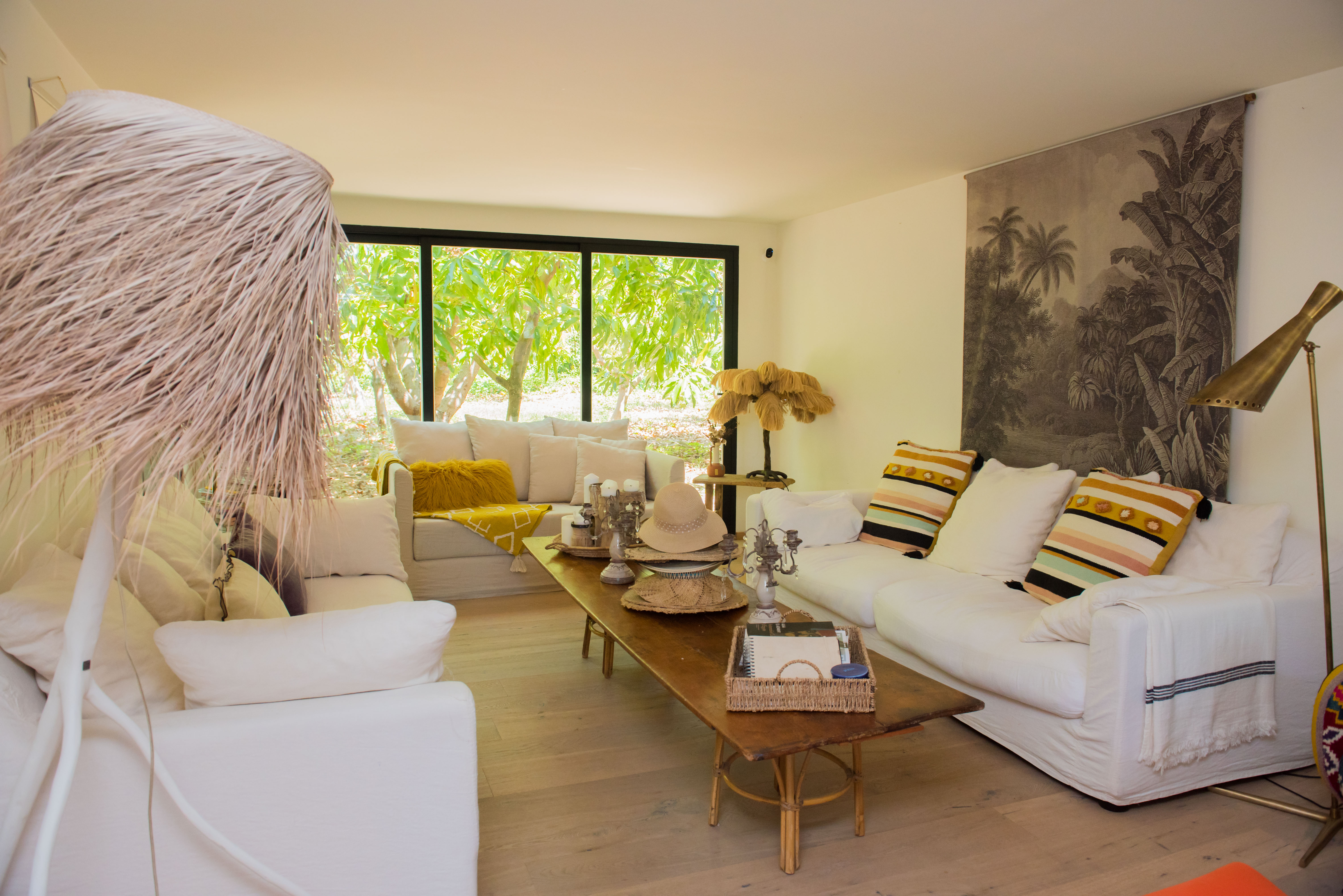 Amplia sala de estar con suelos de madera clara y sofás blancos. Grandes ventanales enmarcan vistas a un exuberante jardín tropical, realzados por una mesa de centro rústica y lámparas decorativas únicas.