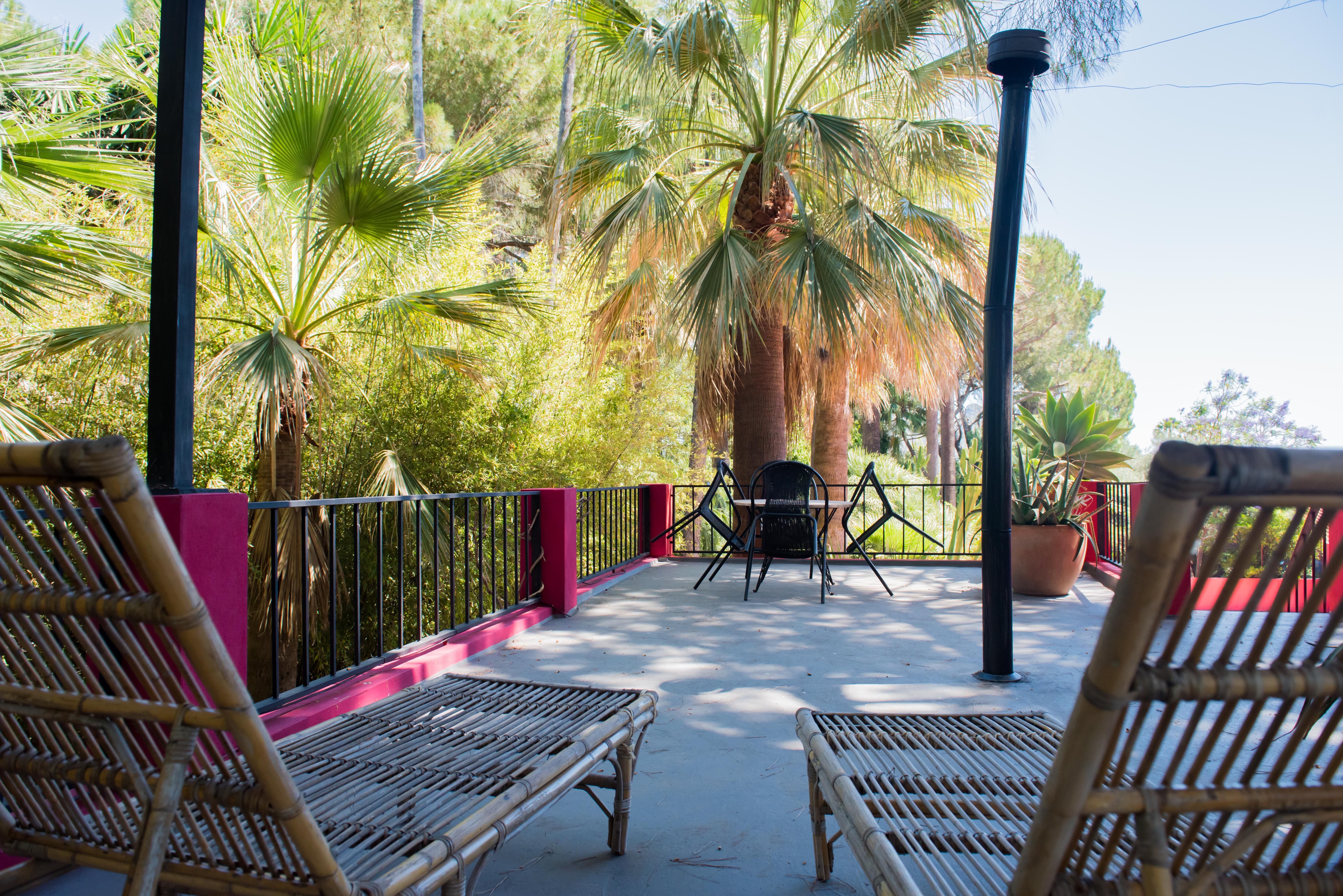 Amplia terraza exterior con dos tumbonas de bambú y un conjunto de mesa y sillas, enmarcada por un exuberante jardín tropical con palmeras y barandales fucsia. El suelo de hormigón recibe luz natural.
