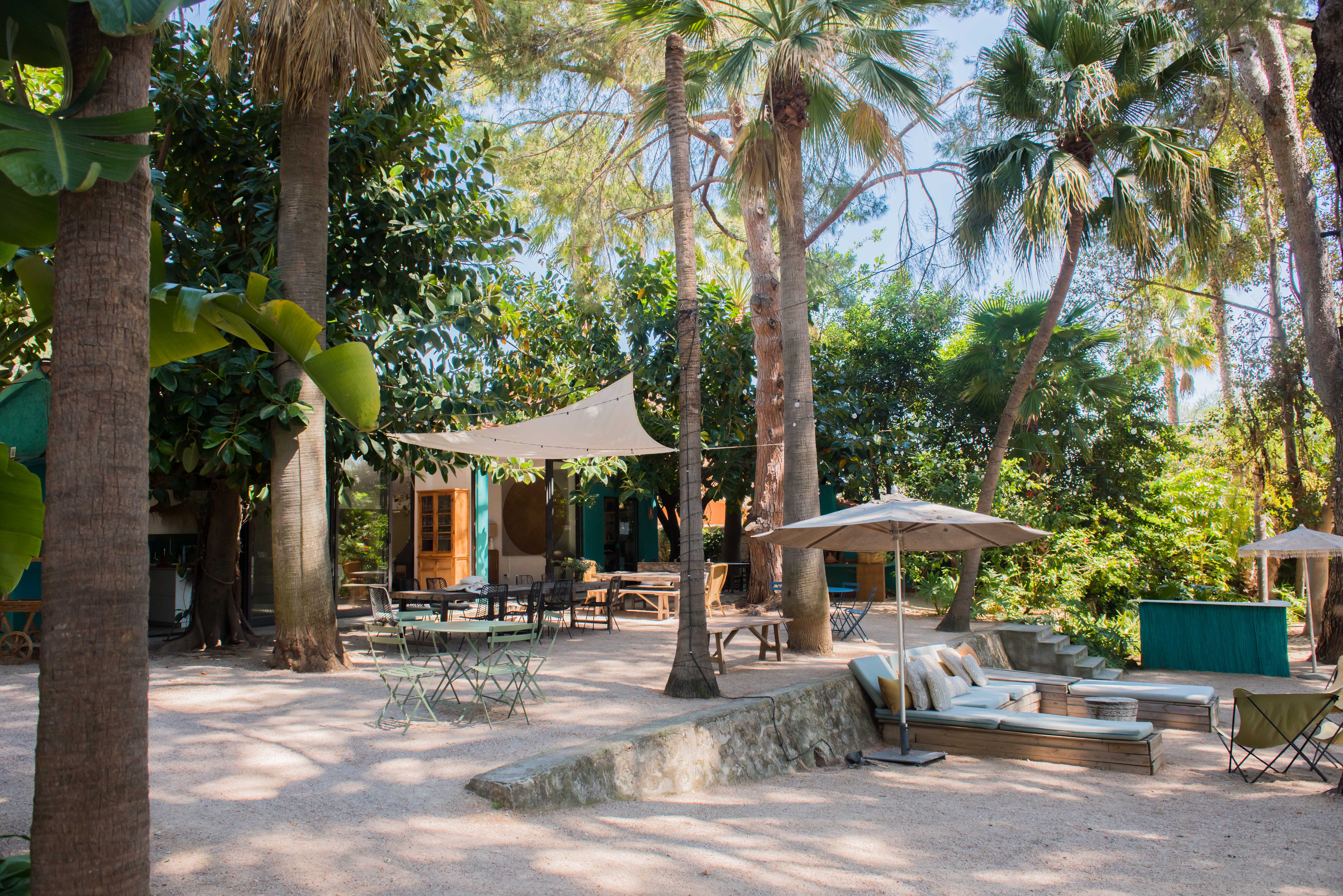 Amplio patio ajardinado tropical con varias zonas de asientos sombreadas sobre grava, incluyendo una gran mesa de comedor bajo un toldo con luces y un área lounge acolchada.