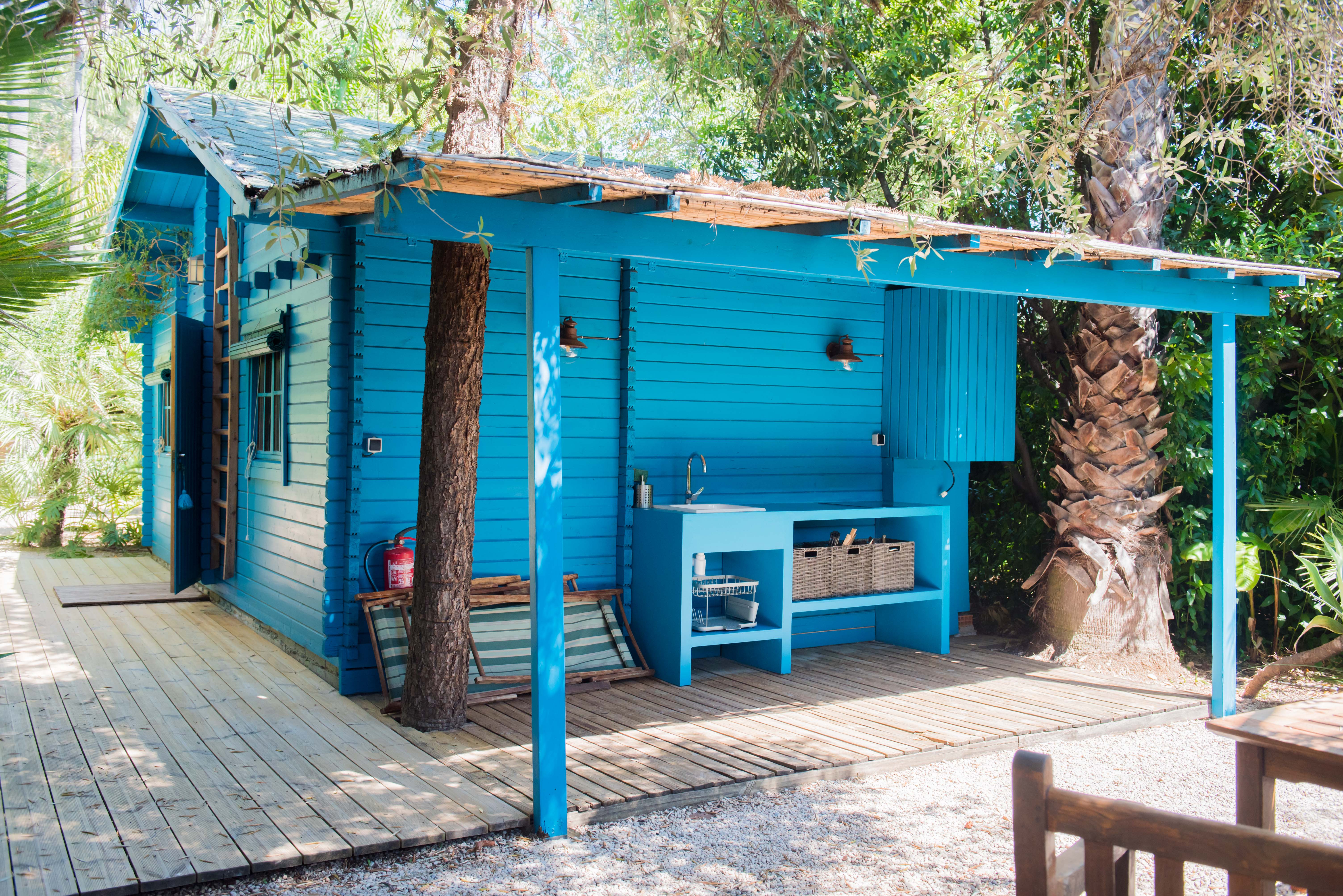 Cabaña de madera azul vibrante con cocina exterior integrada, fregadero y estantes abiertos, sobre una tarima de madera. Árboles tropicales crecen a través de su dosel natural, rodeada de caminos de grava.
