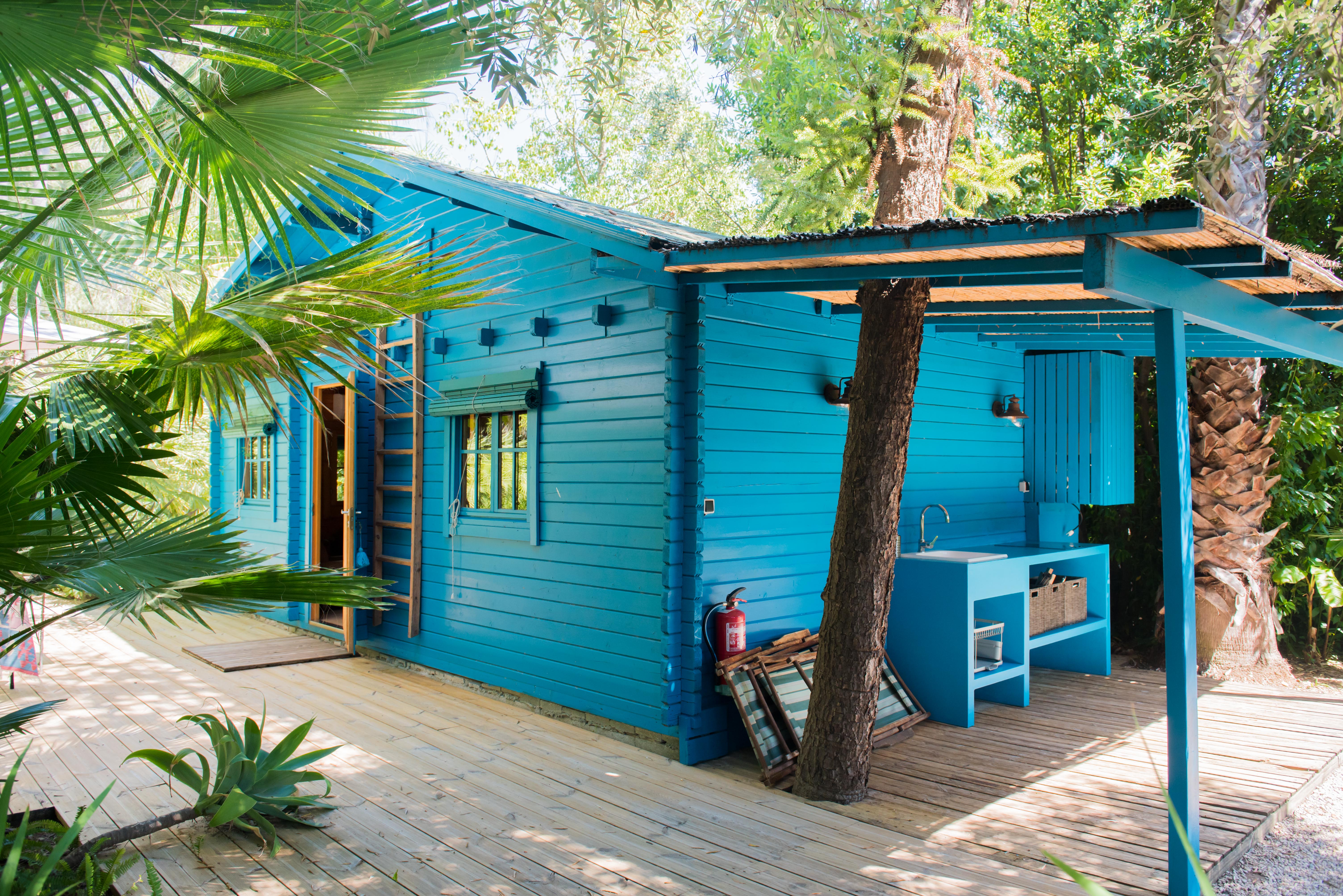 Cabaña de madera azul vibrante con cocina exterior integrada y fregadero, sobre una amplia terraza de madera. Rodeada de exuberante follaje tropical, un tronco de árbol atraviesa el área exterior sombreada.