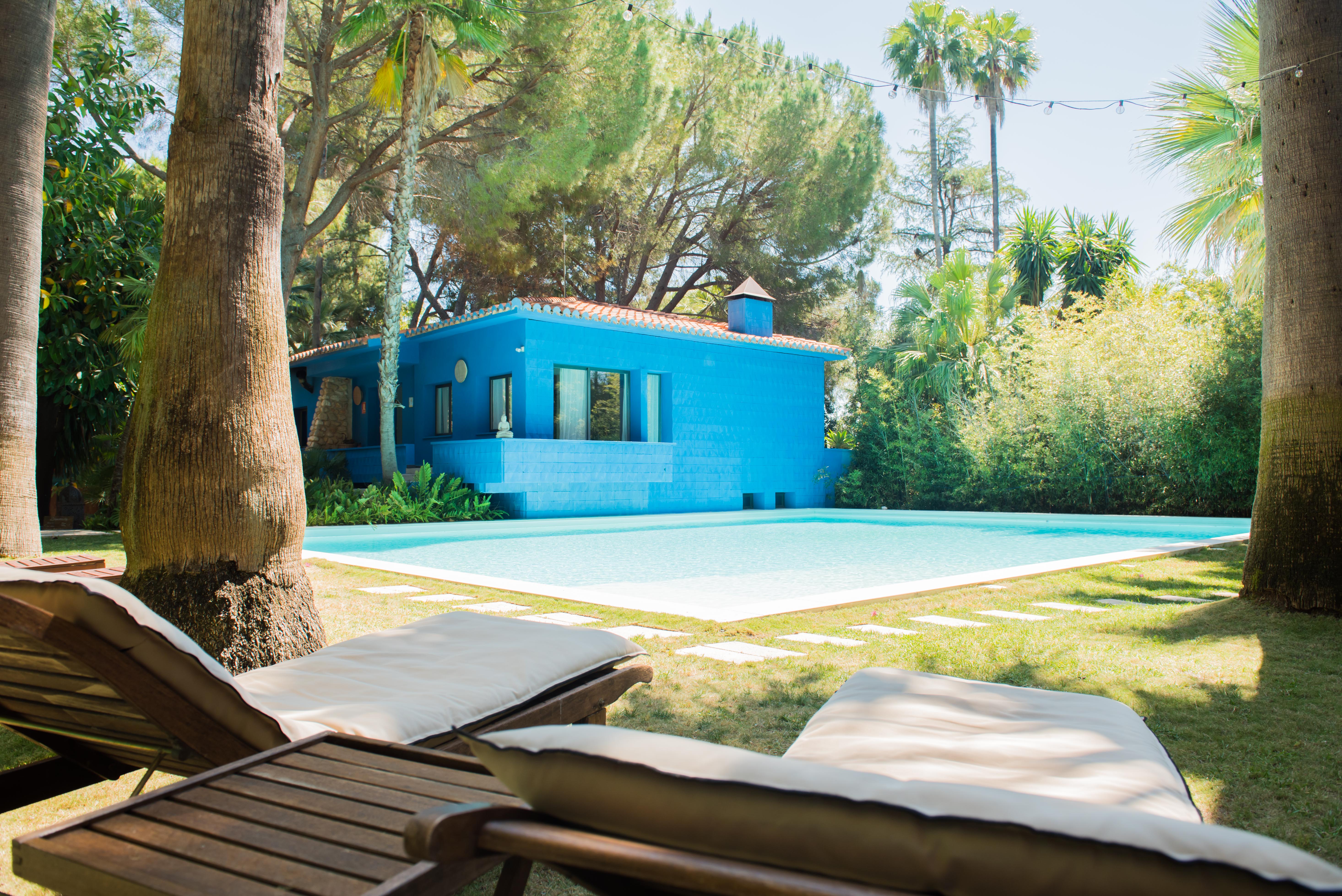 Casa azul vibrante con tejado de terracota, junto a una piscina de agua clara. Dos tumbonas de madera sobre el césped cuidado, enmarcadas por palmeras tropicales y vegetación frondosa bajo luces festivas.