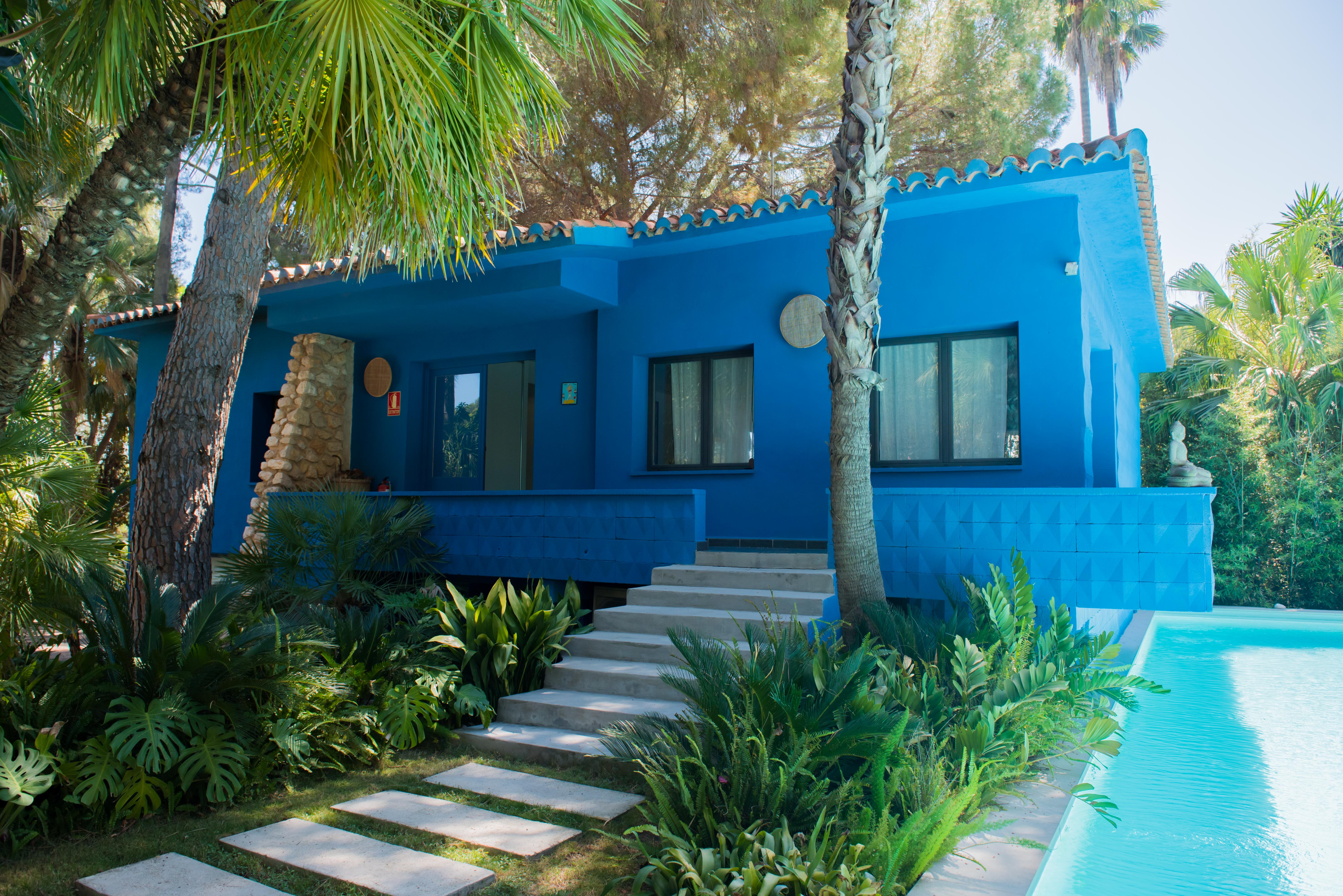 Casa azul vibrante con tejado tradicional, rodeada de exuberantes jardines tropicales y palmeras. Escaleras de hormigón conducen a una terraza elevada con vistas a una serena piscina.