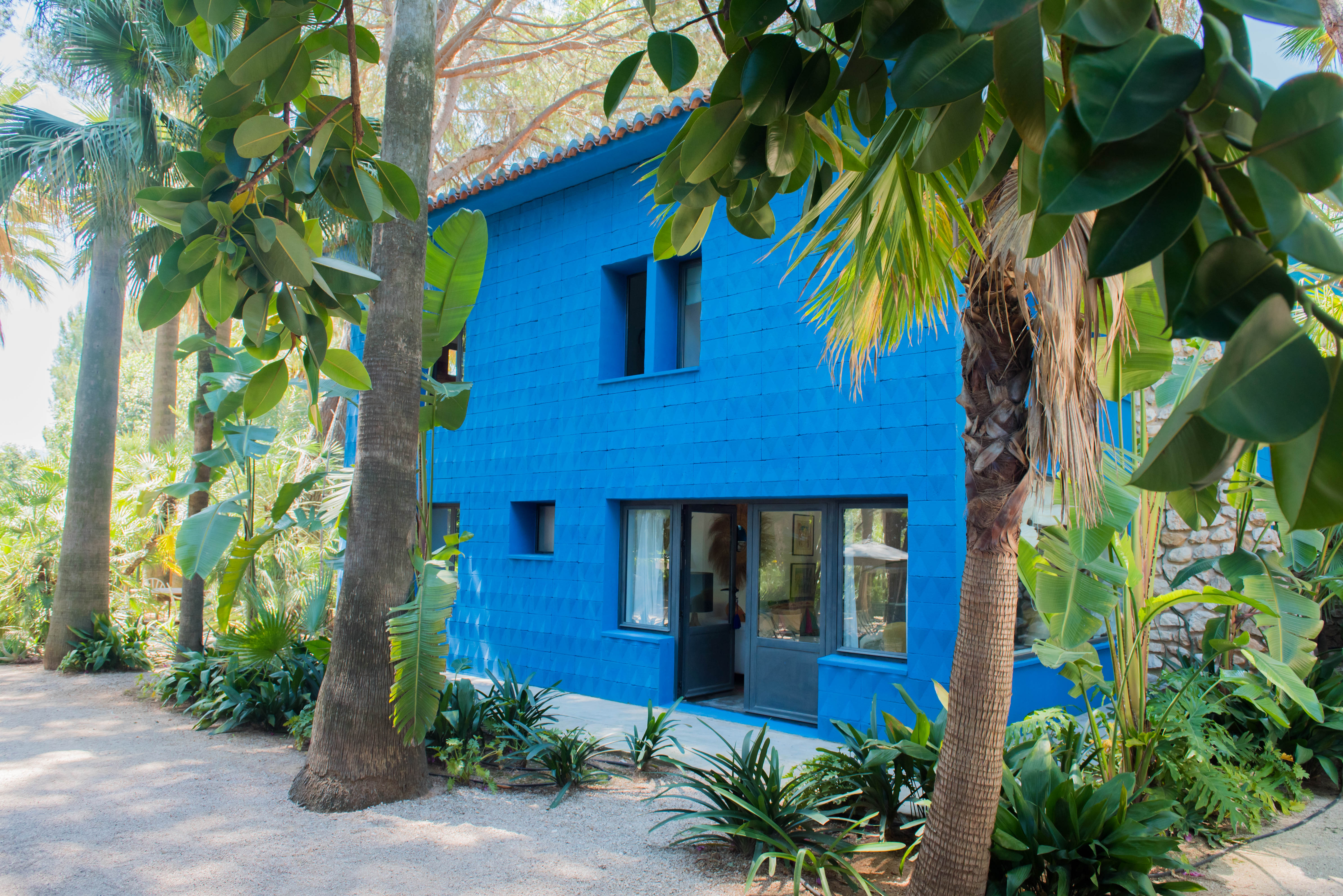 Casa de dos plantas con fachada azul vibrante y textura geométrica, tejado de tejas de terracota, rodeada por un exuberante jardín tropical con palmeras y un sendero de grava.