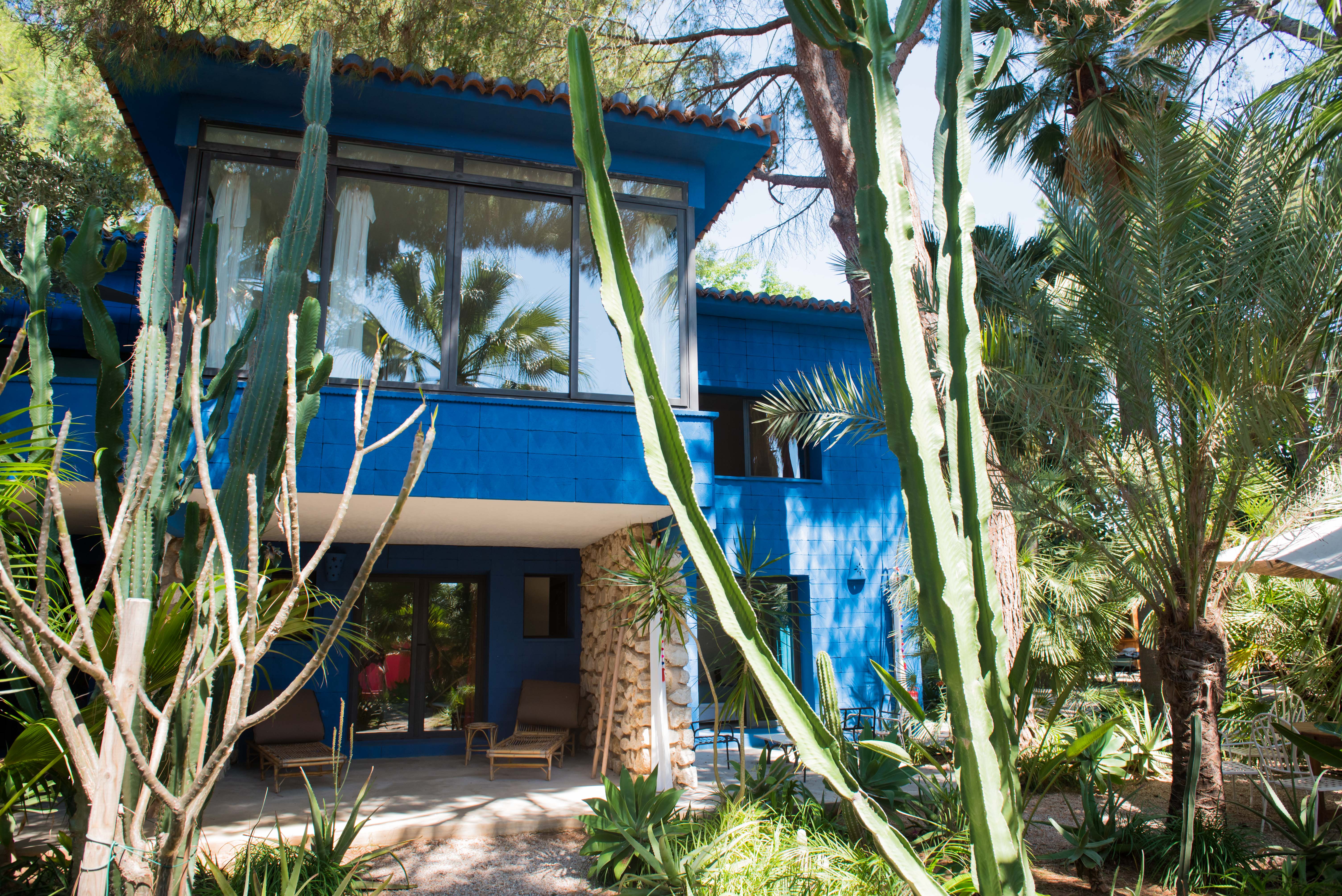 Casa de dos plantas en azul vibrante con patio sombreado en planta baja, tumbonas de bambú y pilares de piedra. El jardín tropical está enmarcado por cactus altos y palmeras.