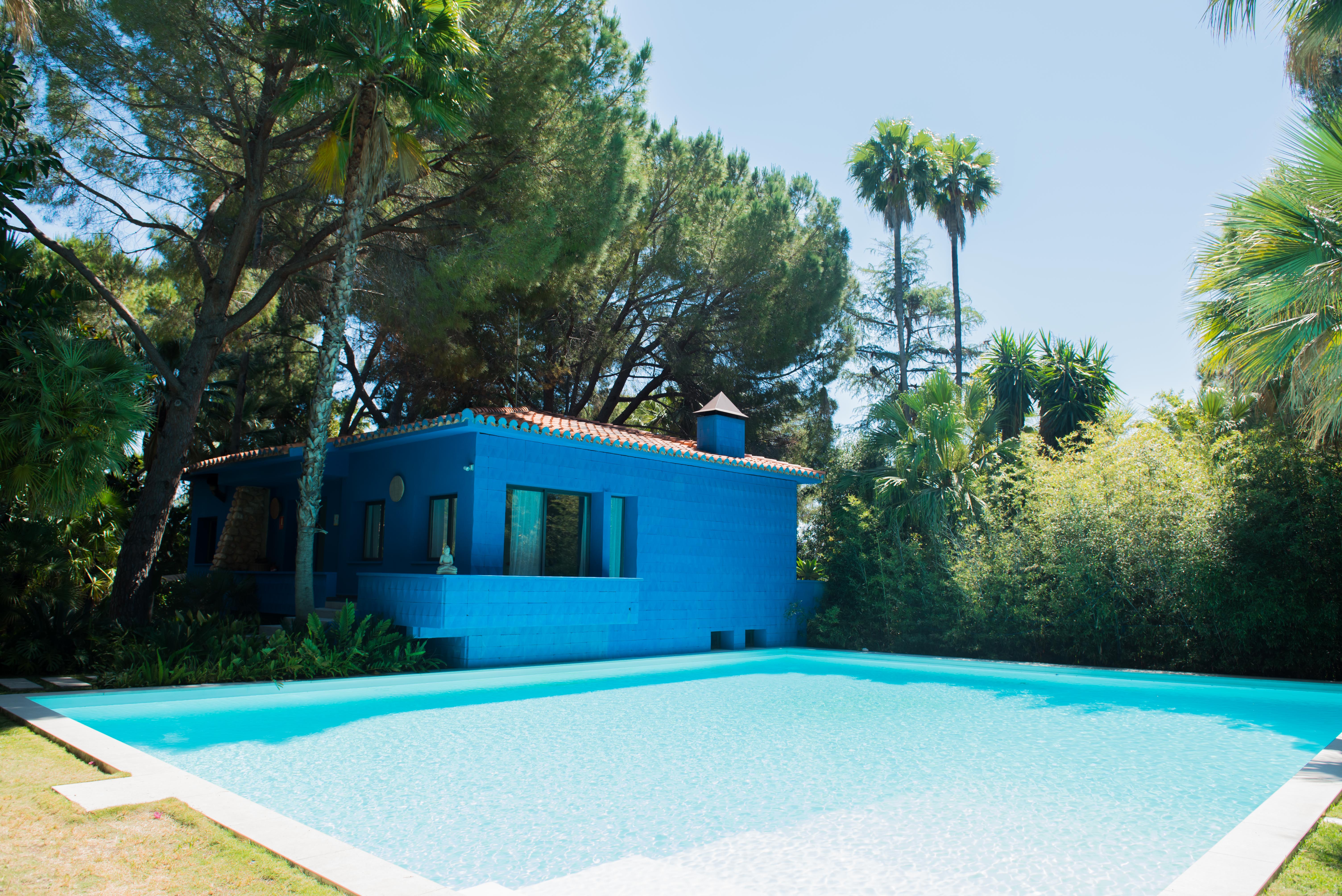 Casa moderna azul con tejado de terracota y una serena piscina con borde claro. Rodeada de exuberante vegetación tropical, incluyendo pinos y palmeras, bajo un cielo despejado.