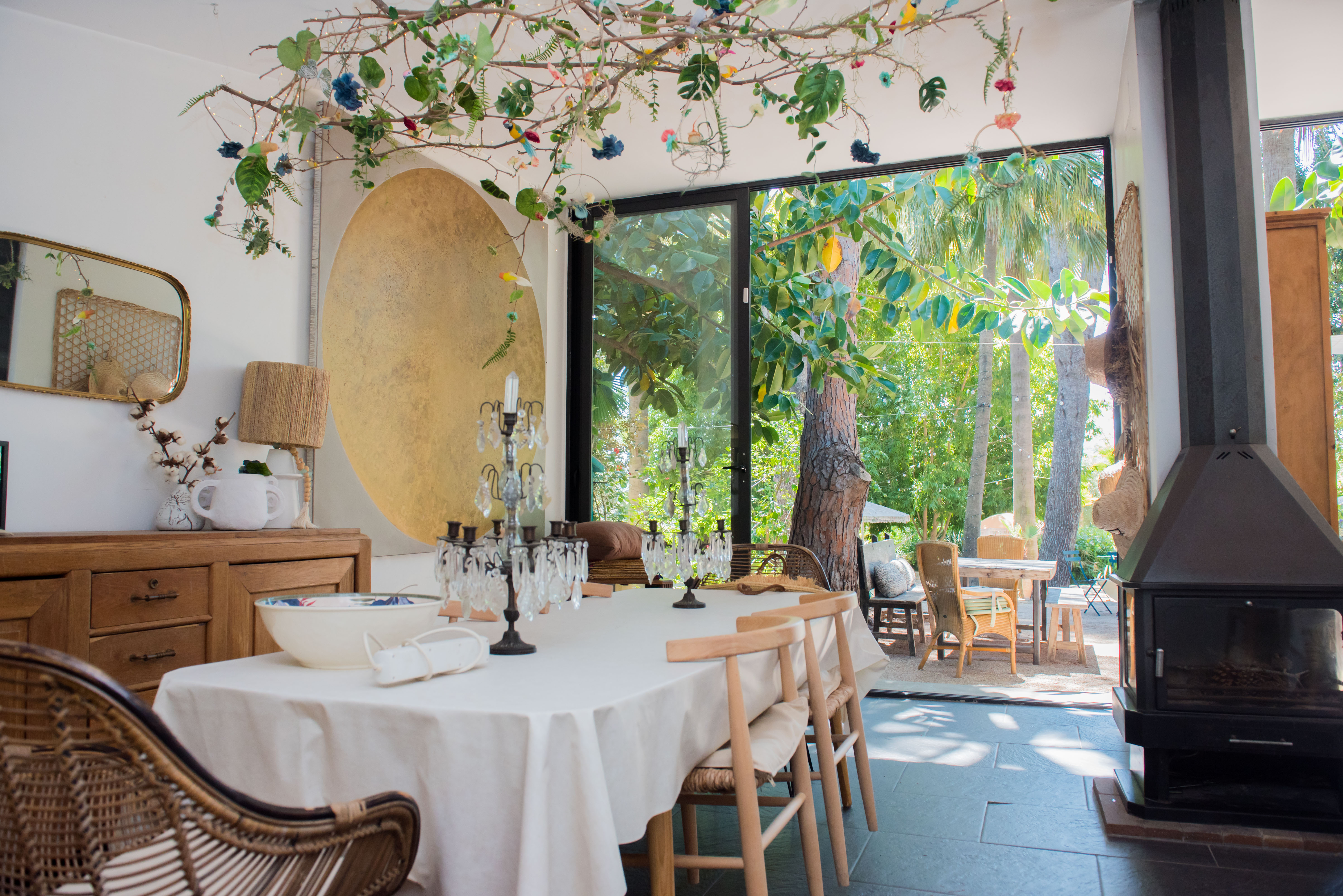Comedor ecléctico con mesa formal y candelabros de cristal, frente a puertas de vidrio con vista a jardín tropical. Una instalación de ramas en el techo con luces y una chimenea moderna completan el espacio.