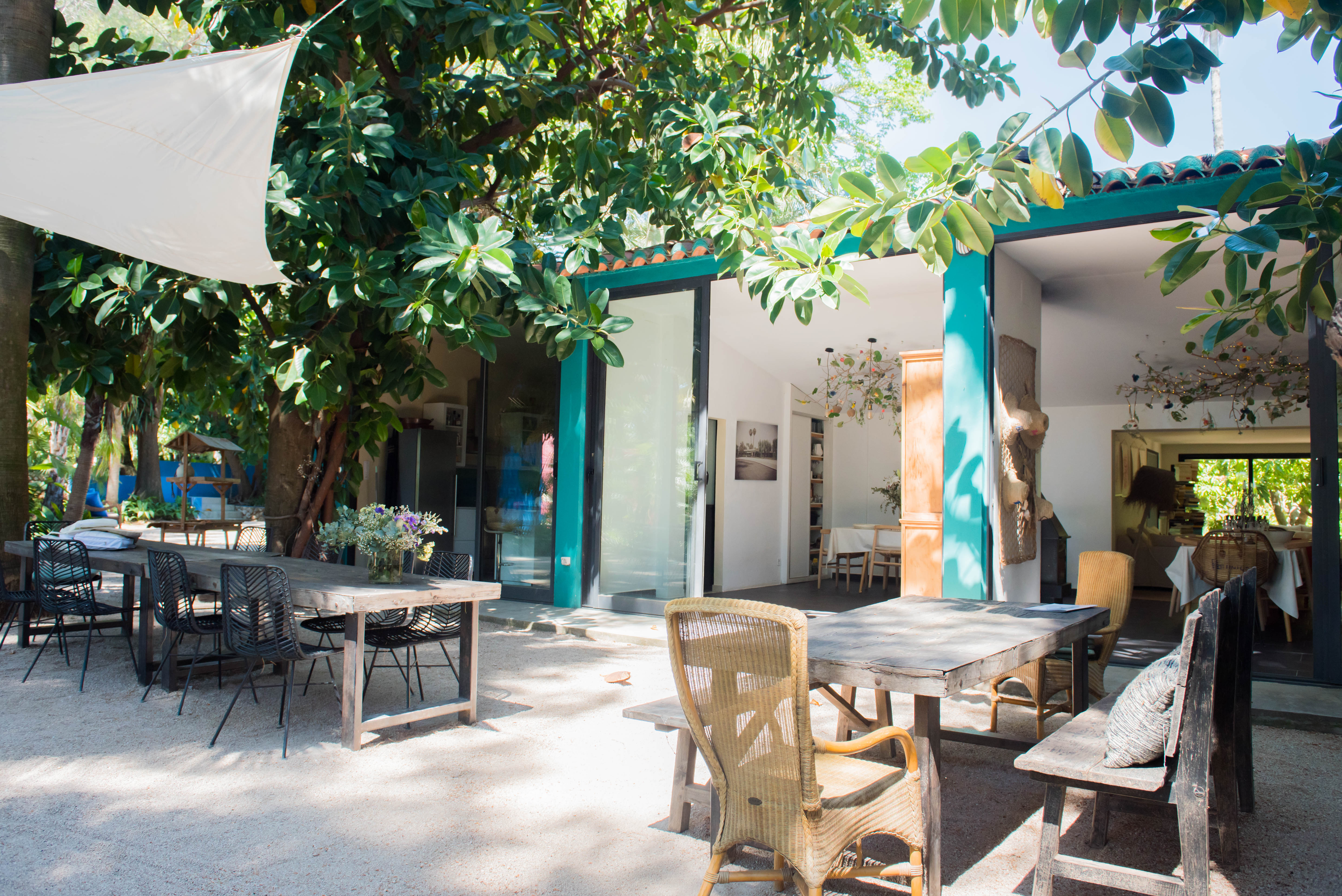 Comedor exterior con mesas de madera y sillas variadas bajo una vela de sombra blanca, enmarcado por exuberante vegetación tropical y conectado al interior por puertas de cristal.