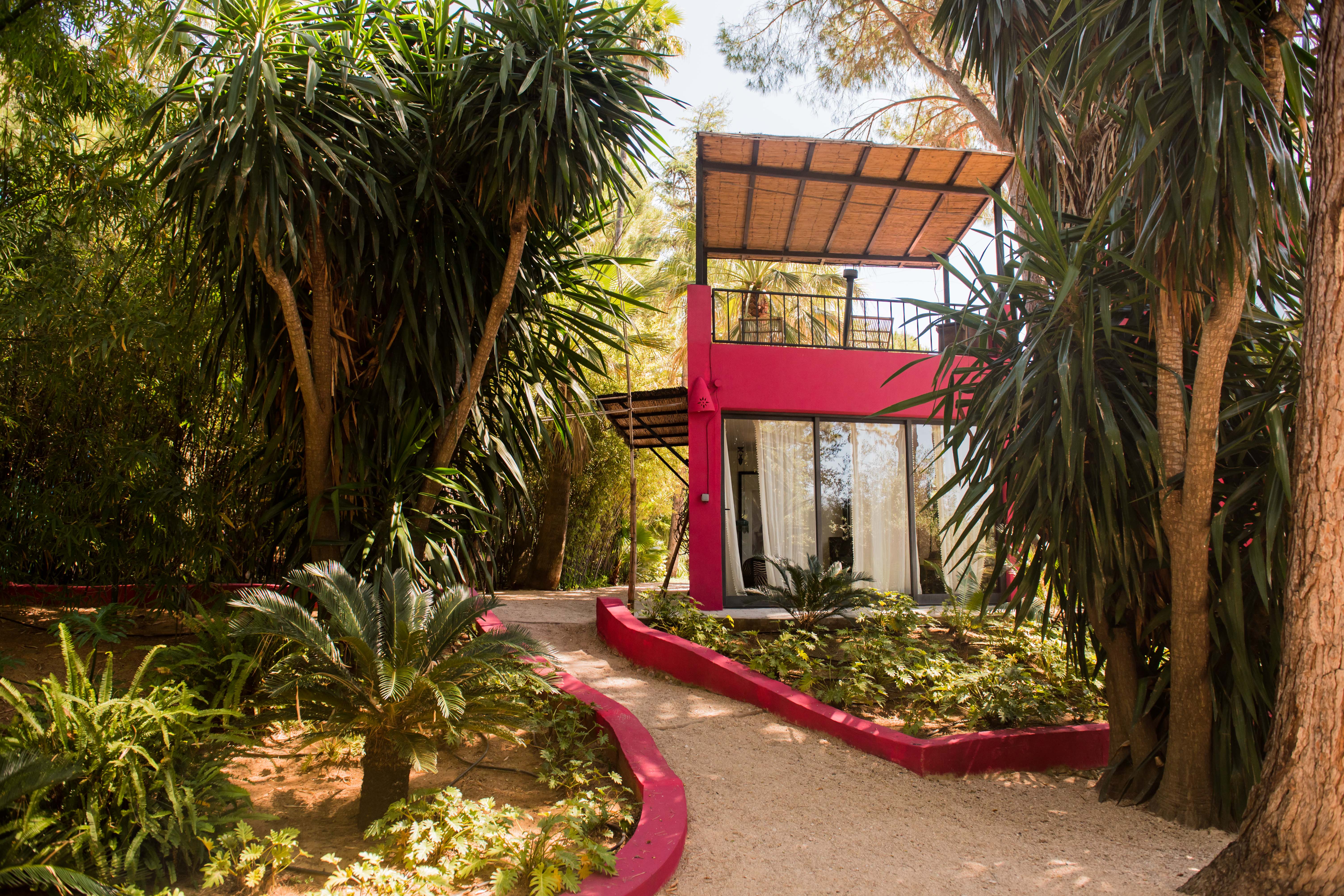 Edificio moderno color fucsia con amplias puertas de cristal, dosel de material natural tejido y balcón superior con asientos de ratán. Un sendero de grava serpentea entre la exuberante vegetación tropical.