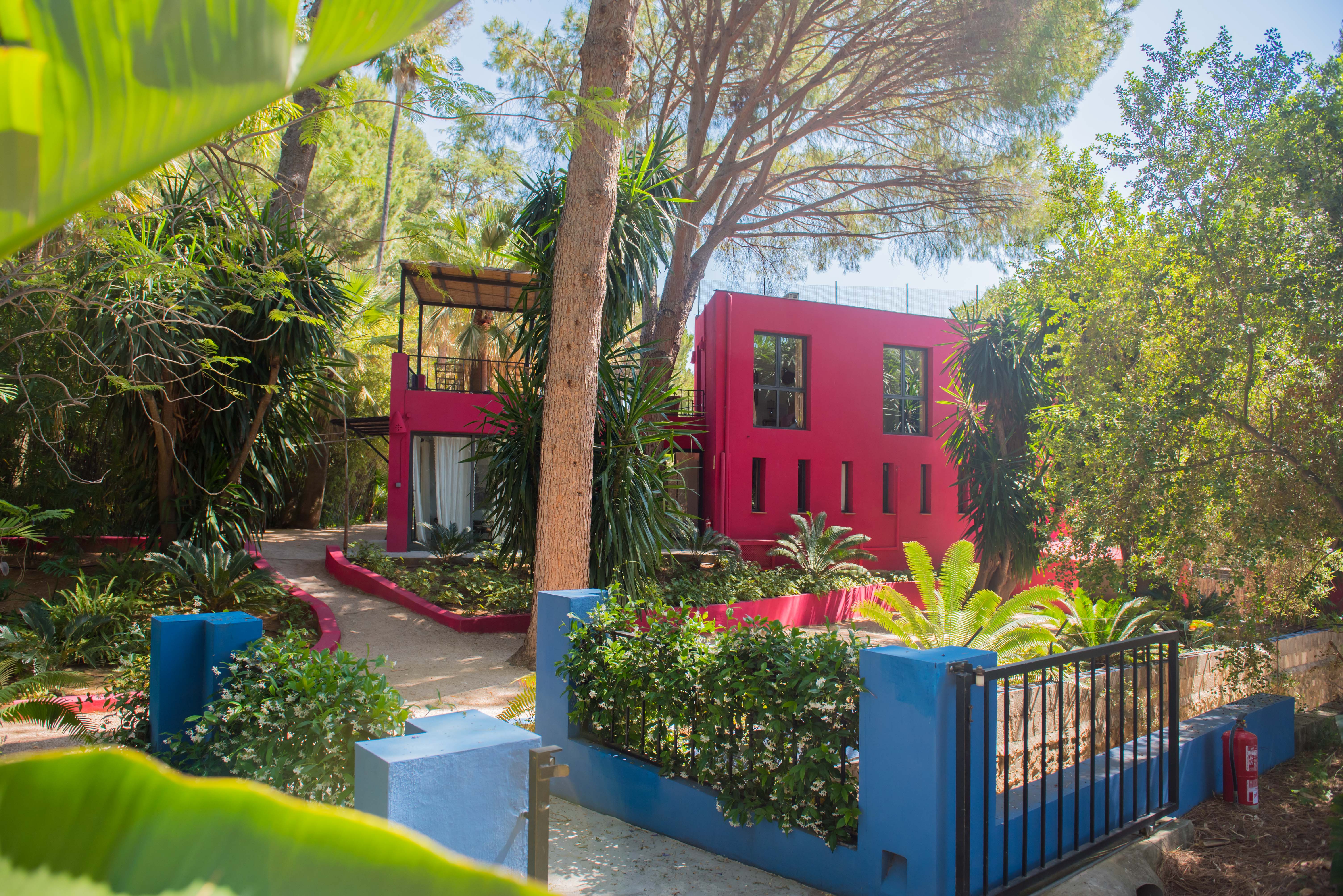 Edificio moderno de color fucsia vibrante con ventanas variadas, enclavado en un denso jardín tropical con árboles altos y senderos serpenteantes. Un muro bajo azul con arbustos en flor enmarca el primer plano.