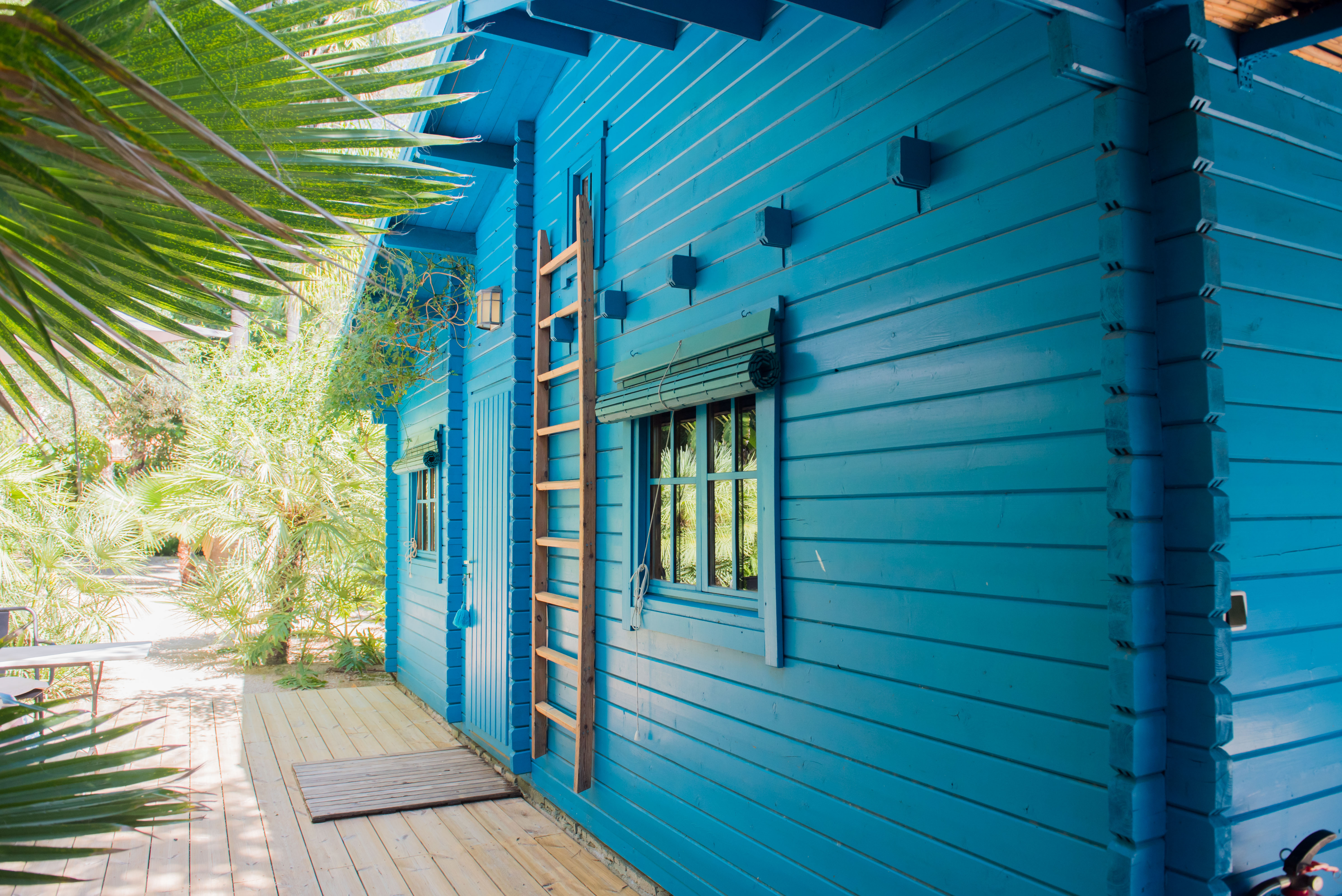Exterior de cabaña de madera azul con escalera decorativa y ventana con persiana de bambú. Una terraza de madera clara se integra en un exuberante jardín tropical.