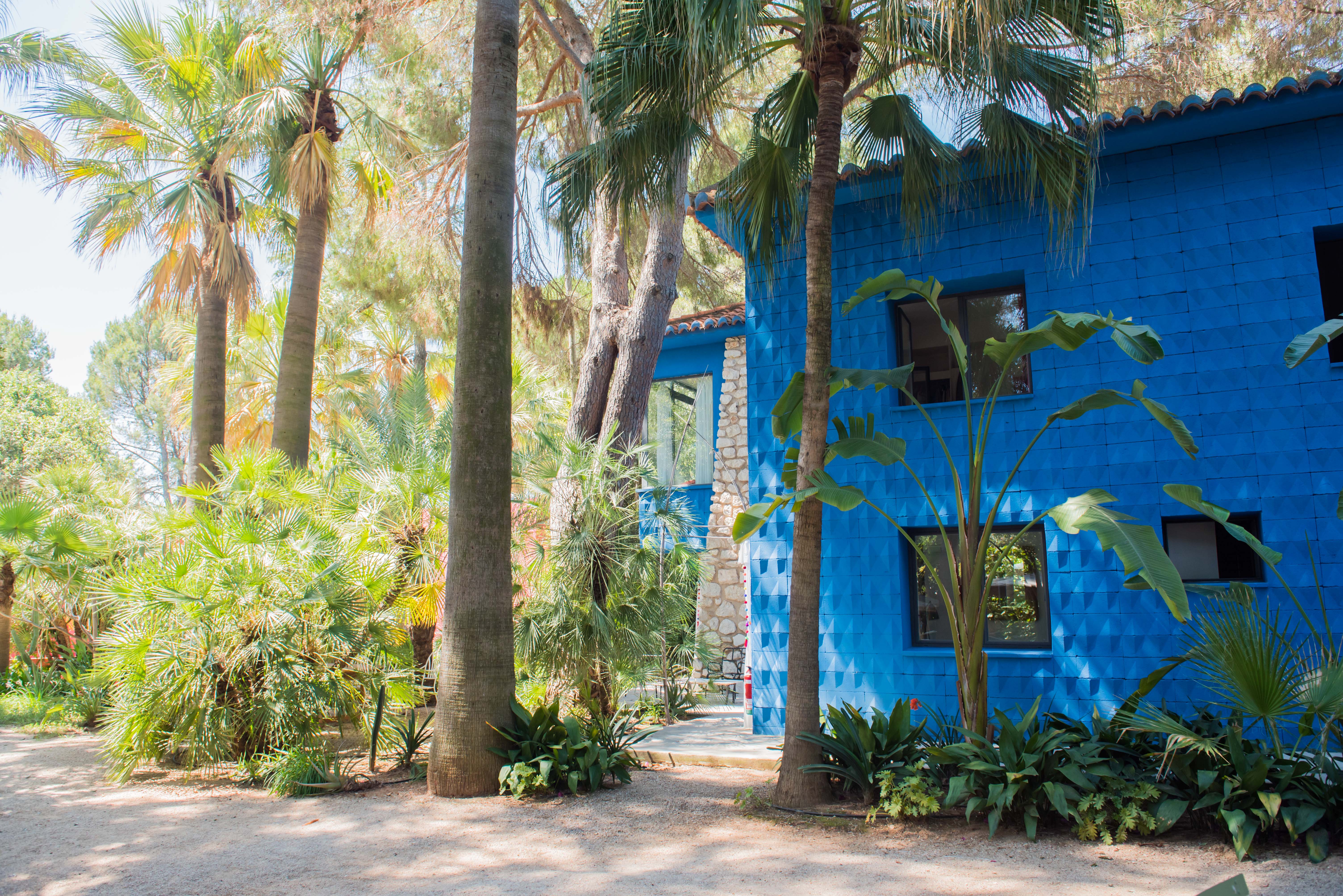 Fachada azul vibrante y texturizada de un edificio, enmarcada por exuberante vegetación tropical. Ventanas oscuras y un techo de tejas de terracota complementan la sección de piedra vista.