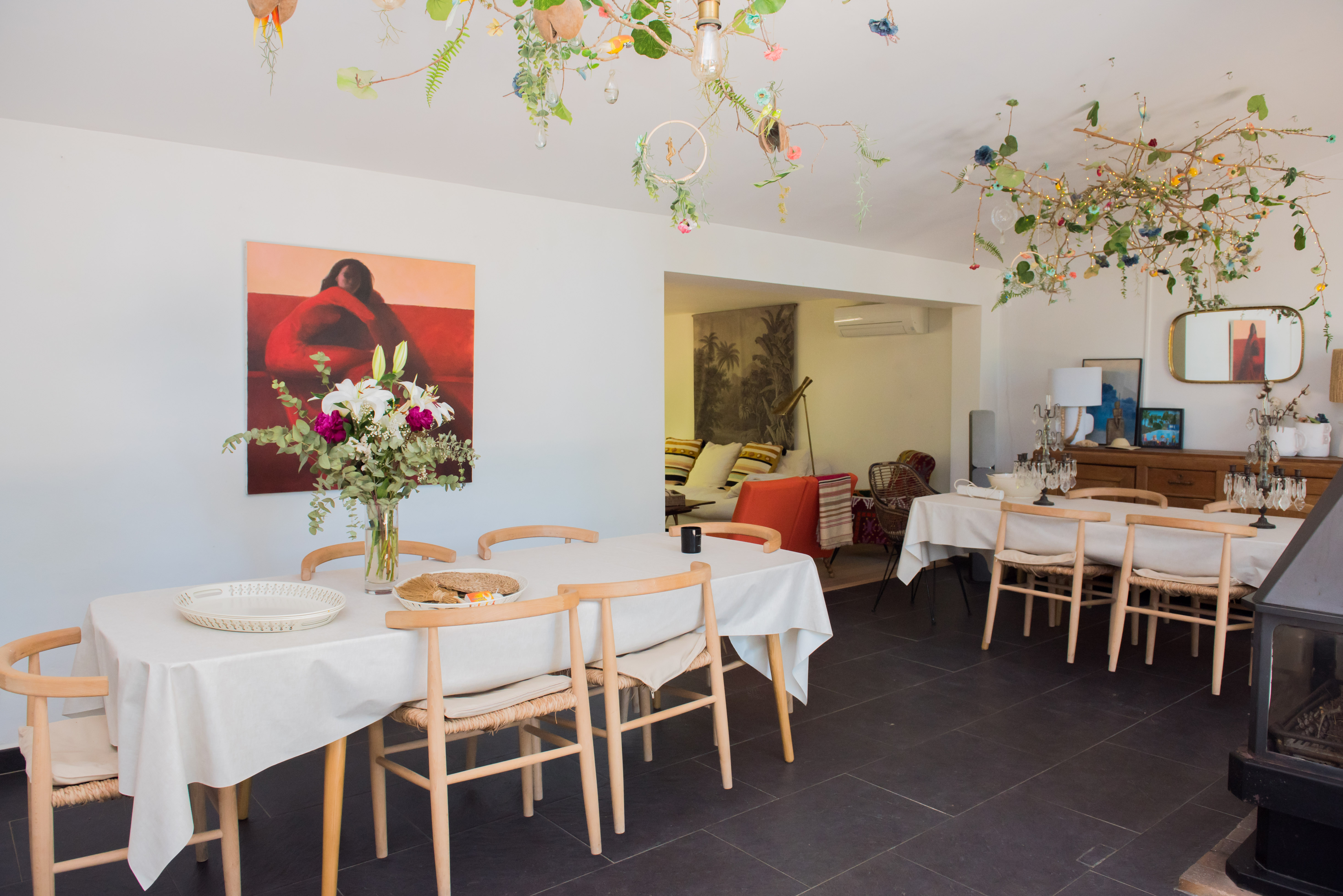 Luminoso comedor con mesa larga, sillas de madera clara y arreglo floral fresco. El techo exhibe una instalación natural única con ramas, flores y luces sutiles, sobre un suelo de baldosas oscuras.