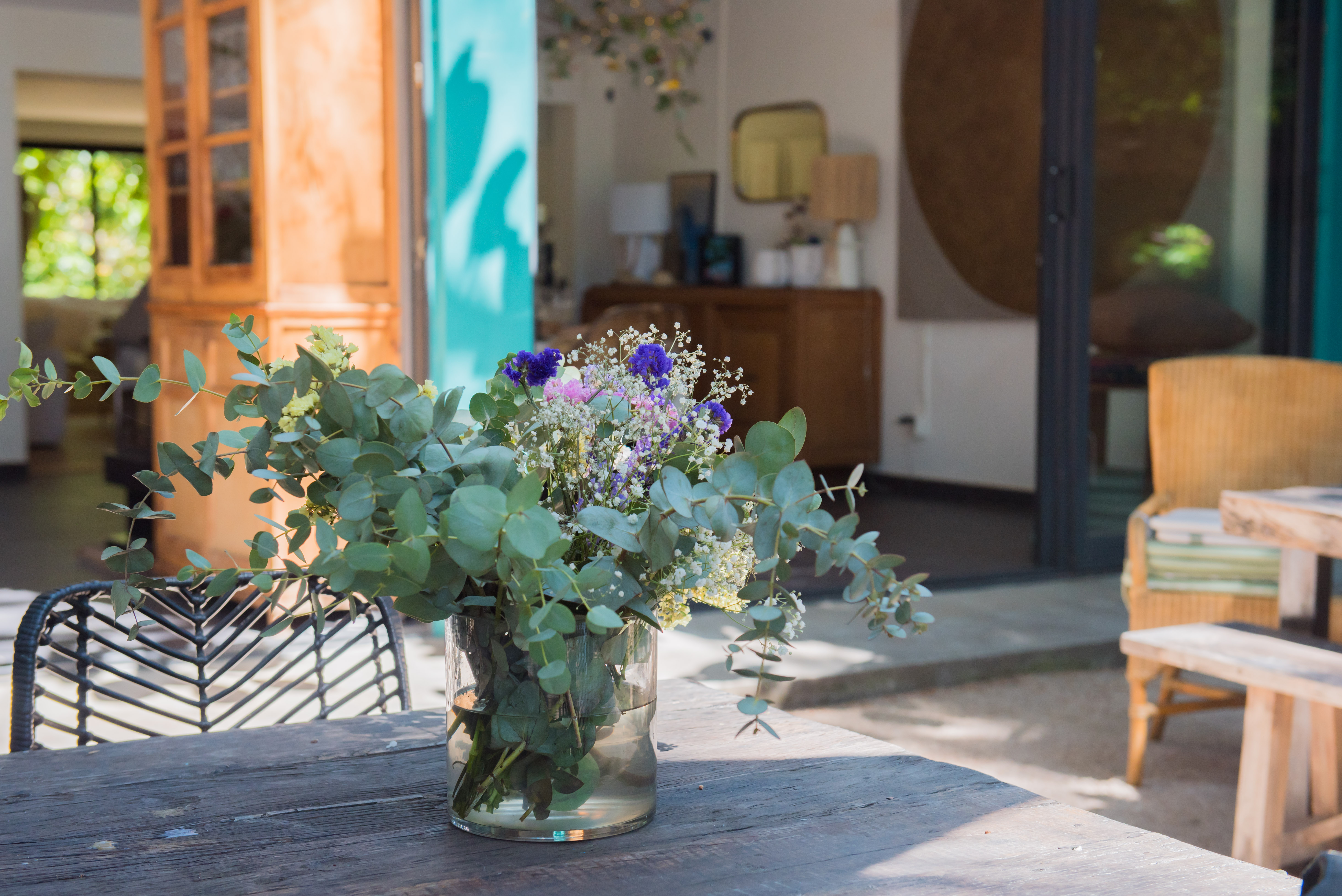 Mesa exterior rústica de madera con un fresco arreglo floral de eucalipto, enmarcada por una puerta turquesa abierta que conecta con un interior luminoso y un jardín exuberante.