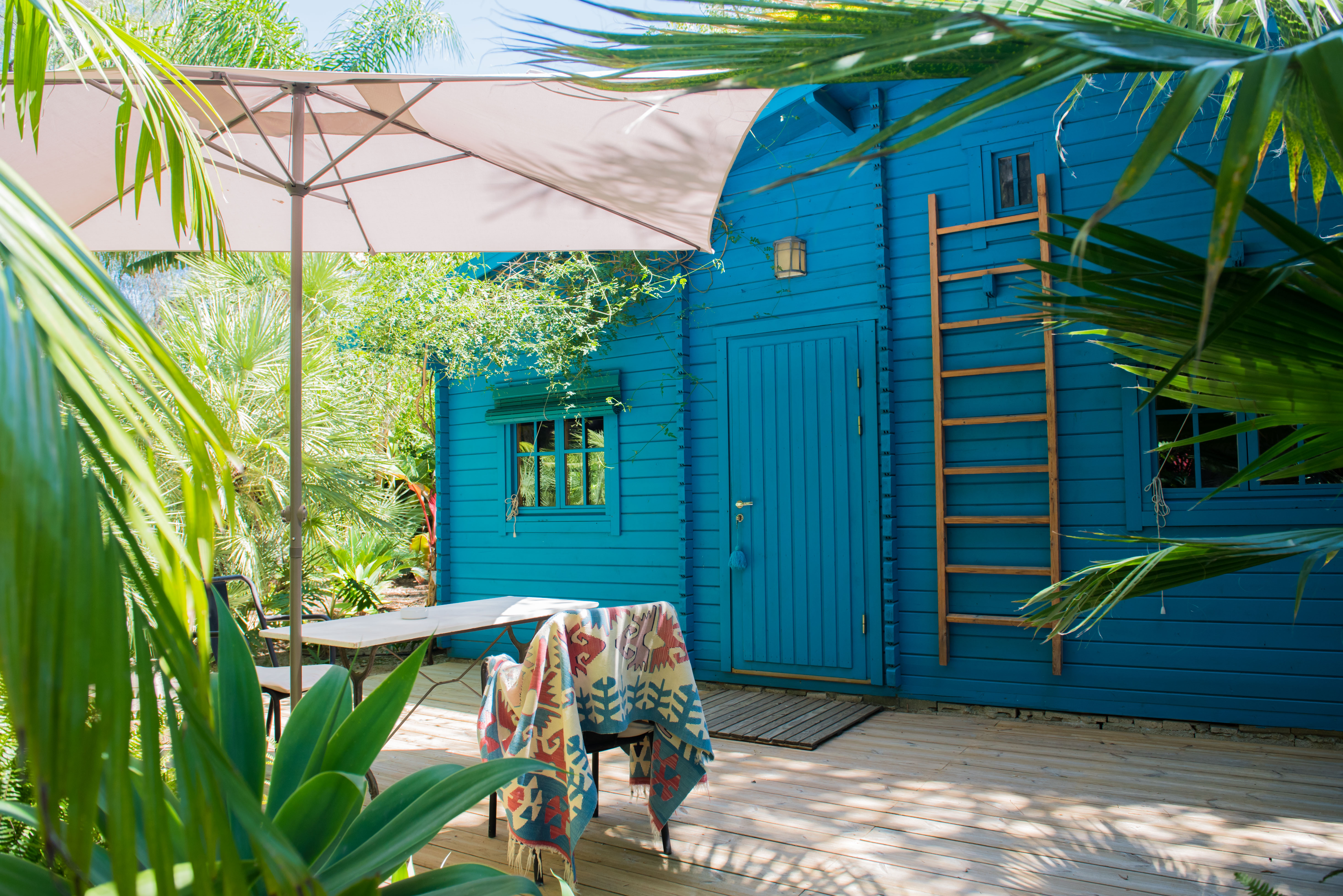 Terraza de madera con sombrilla y mobiliario exterior, destacando una vibrante cabaña azul con escalera de madera, inmersa en un exuberante jardín tropical.