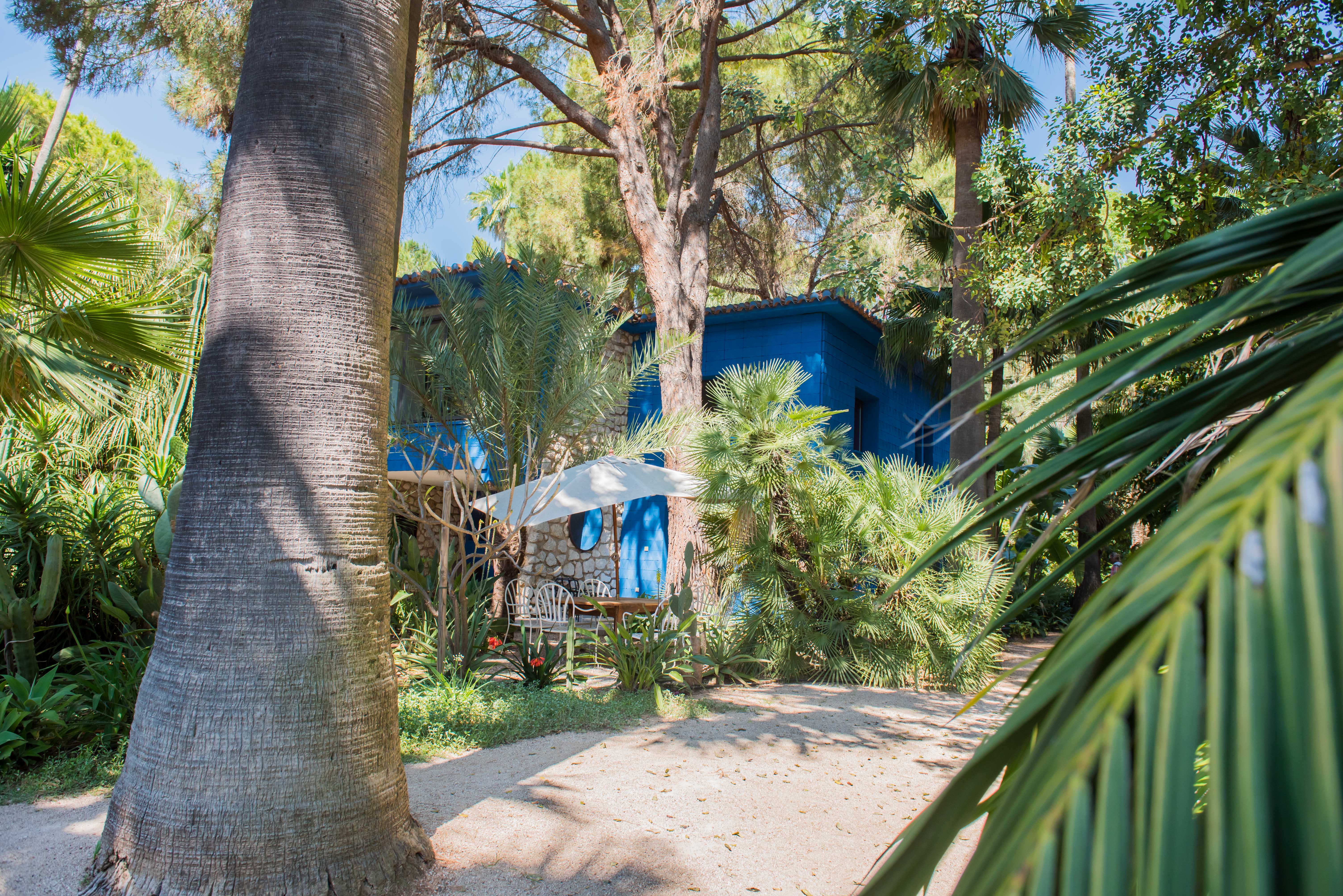 Vista exterior de una casa azul vibrante con detalles de piedra, rodeada por un exuberante jardín tropical. Un área de asientos al aire libre con sillas blancas y sombrilla se ve en un sendero de grava.