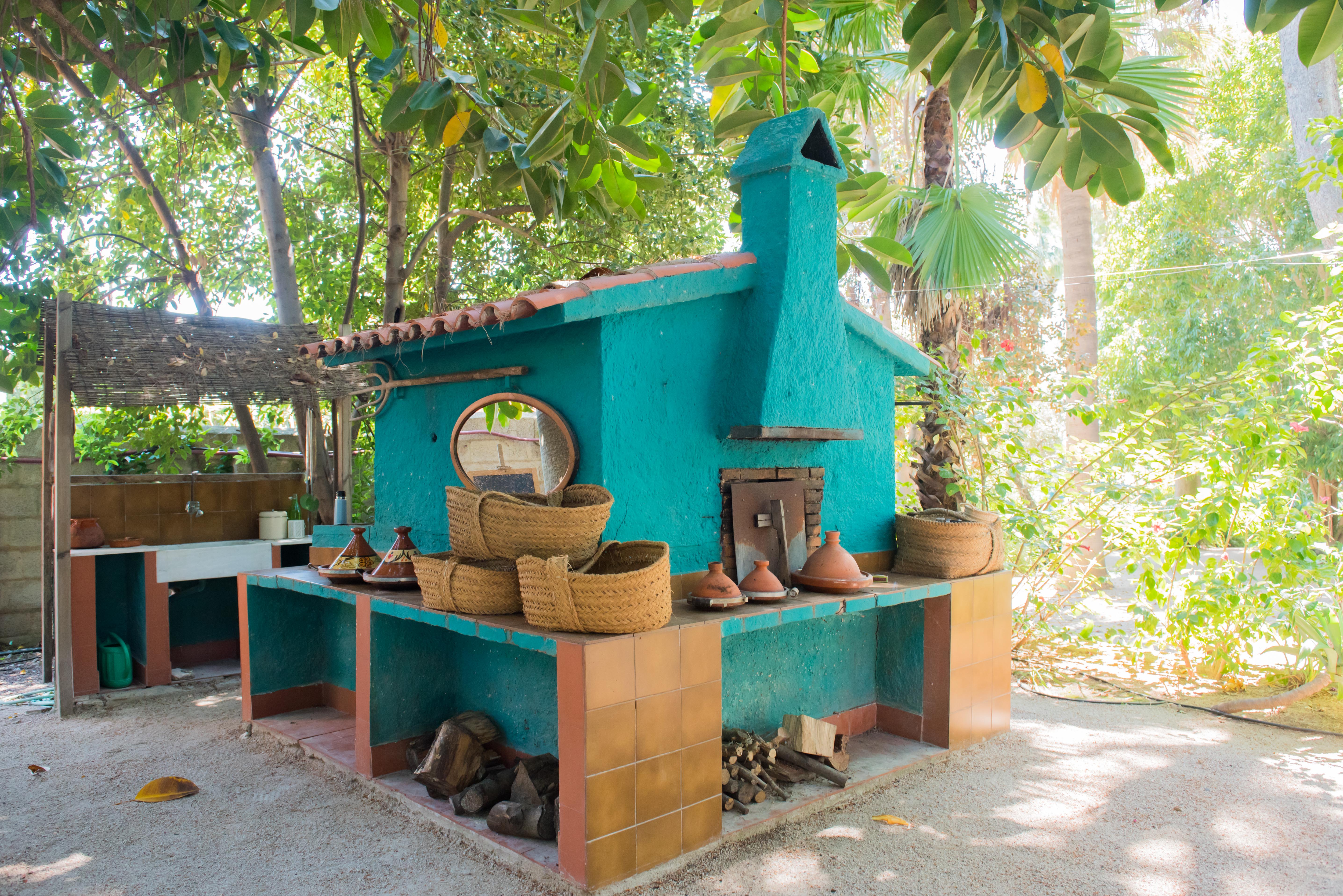 Zona de cocción exterior con horno turquesa y chimenea, encimeras de azulejos terracota y leña almacenada, enmarcada por exuberante vegetación tropical.