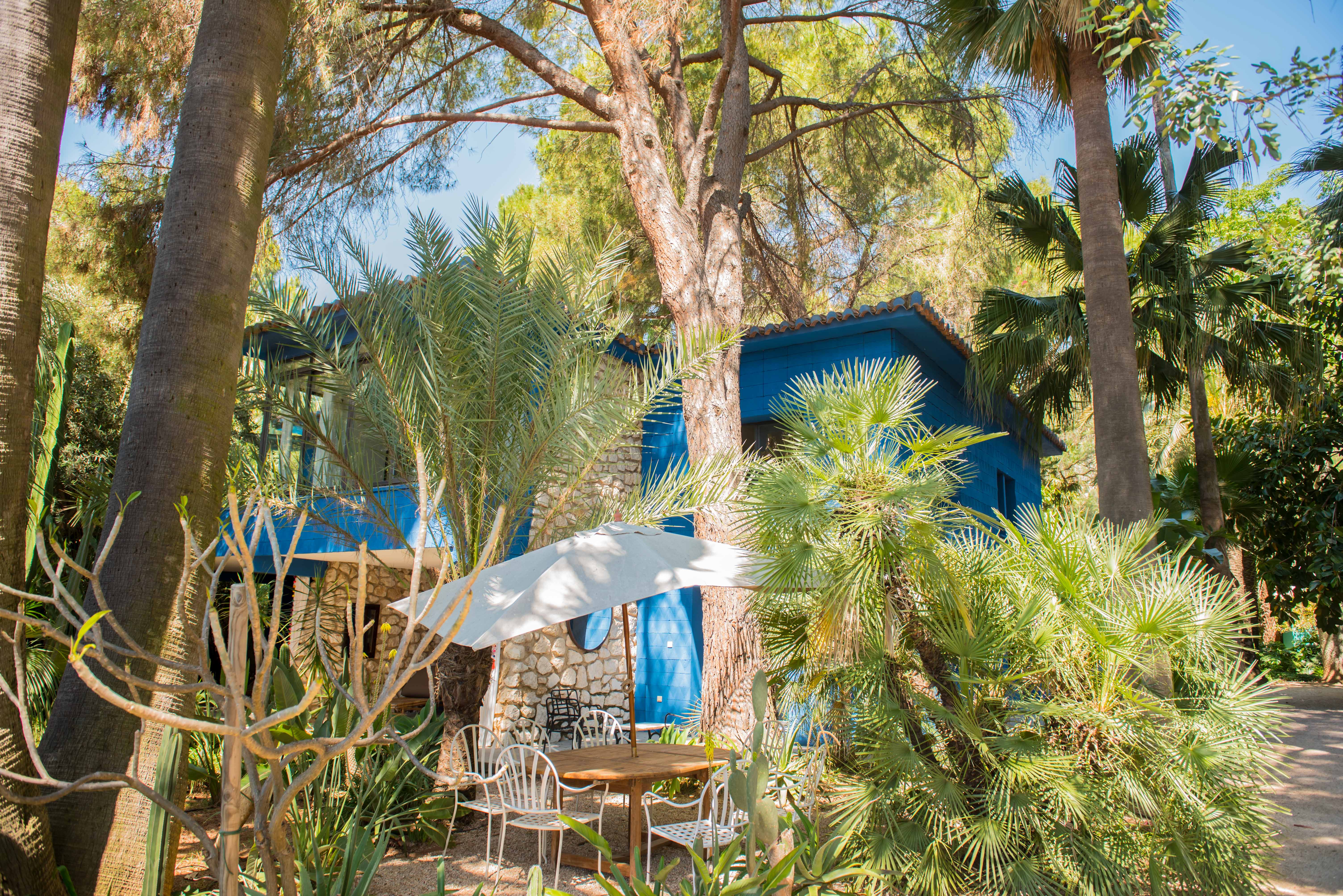 Zona de comedor exterior con mesa de madera y sillas de metal blanco bajo una sombrilla, rodeada de exuberante vegetación tropical. Al fondo, una casa azul vibrante con fachada de piedra.