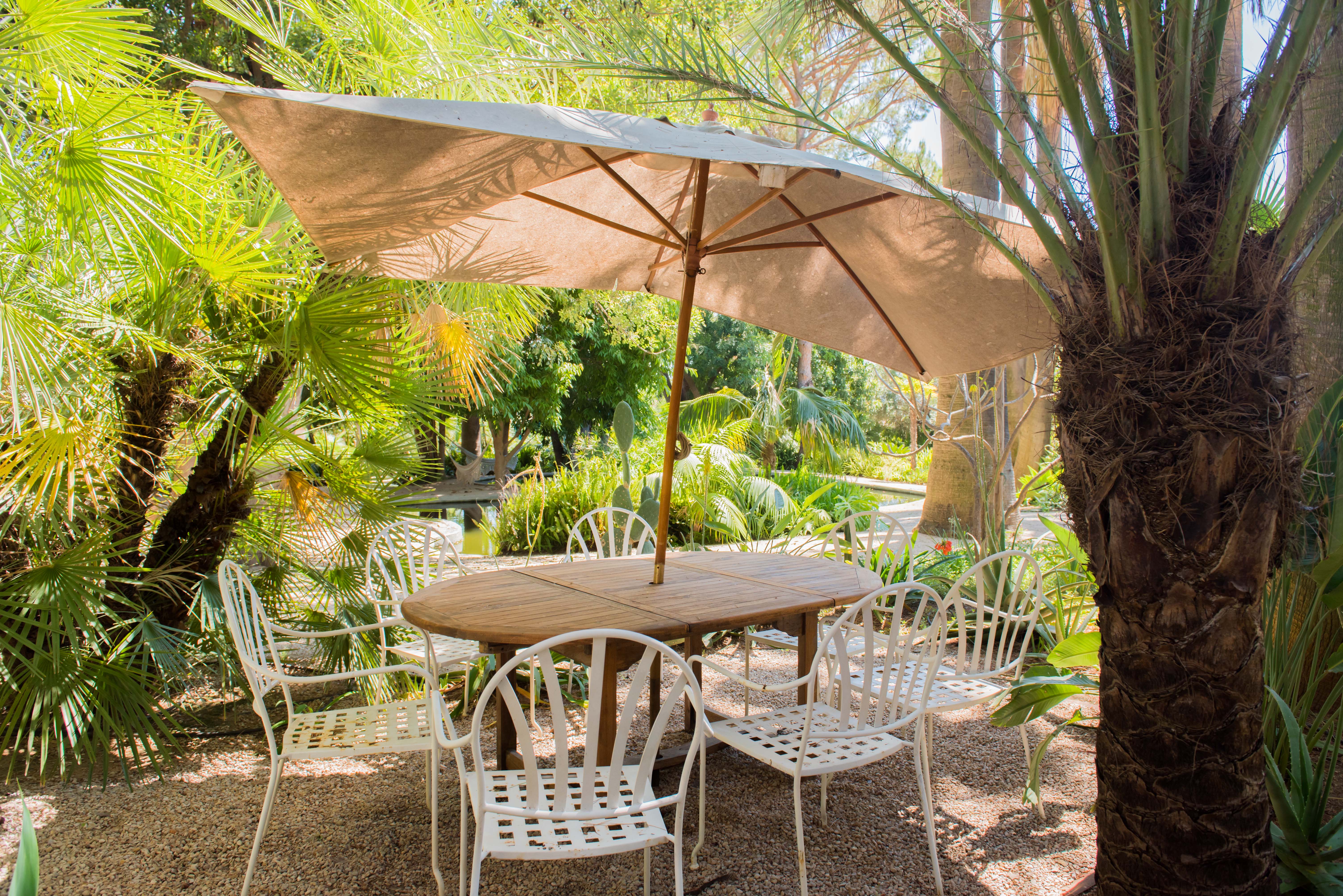 Zona de comedor exterior con mesa redonda de madera y seis sillas de metal blanco bajo una gran sombrilla beige. Rodeado de exuberantes palmeras tropicales y suelo de grava, con un estanque visible al fondo.