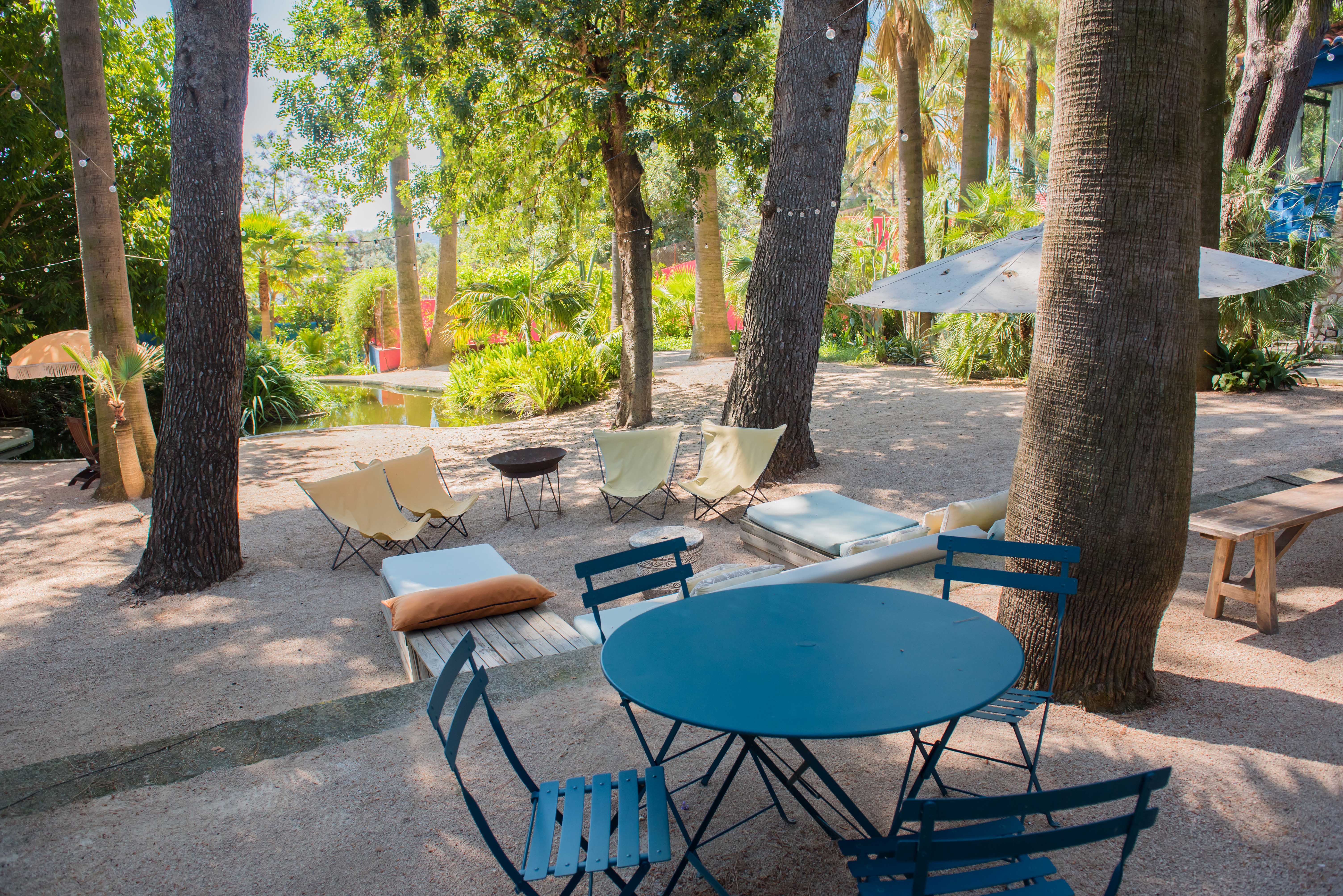 Zona de estar exterior con mesa bistro azul y sillas de tela sobre grava, sombreada por árboles con luces de guirnalda, junto a un estanque.