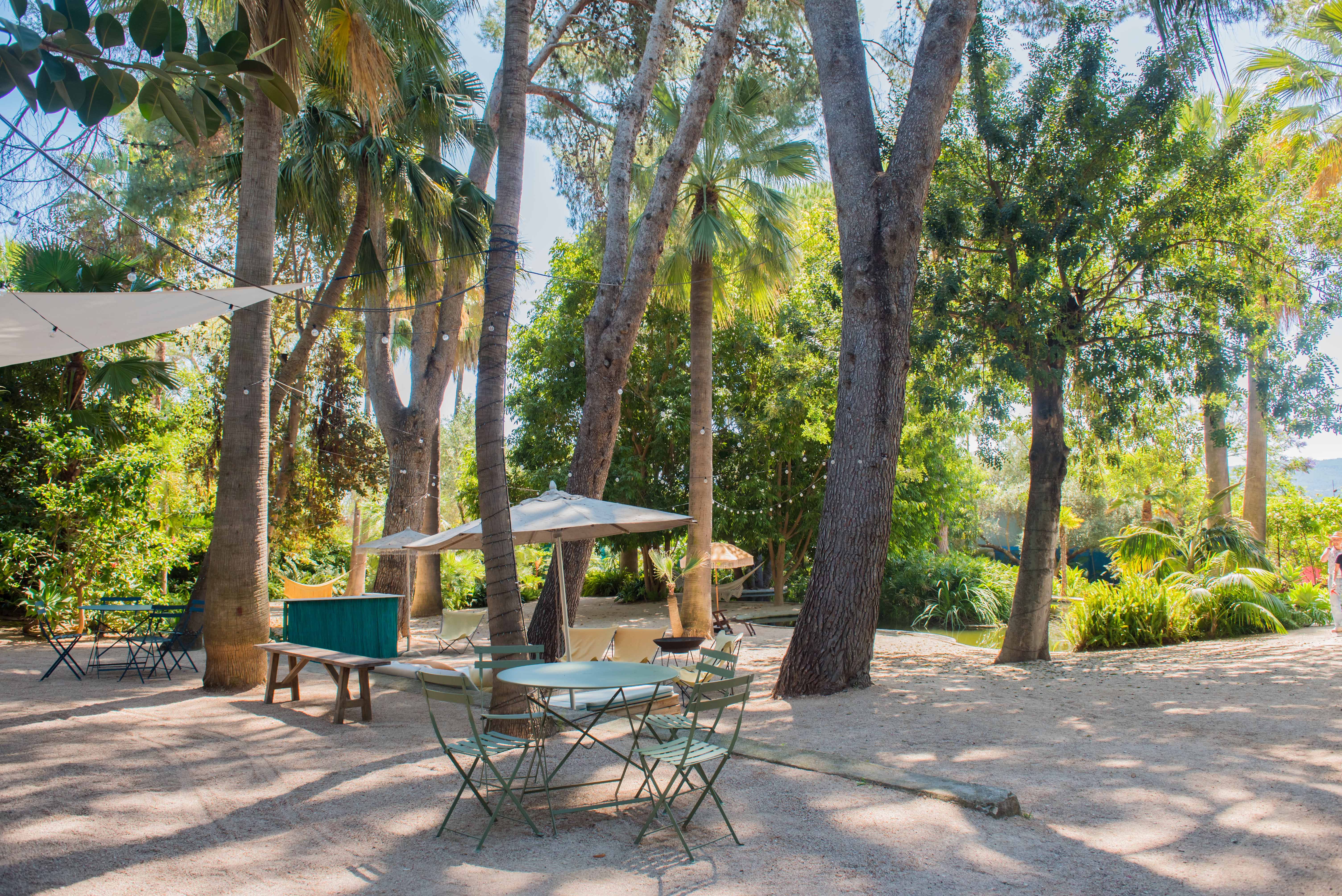 Zona de estar exterior con mesas y sillas de metal verde sobre suelo de grava. Grandes palmeras y árboles maduros ofrecen sombra, complementados por sombrillas y luces colgantes, creando un ambiente tropical.