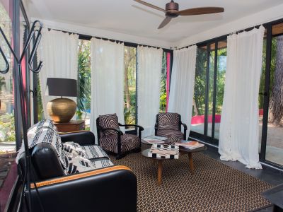 Acogedora sala de estar con amplios ventanales que enmarcan vistas a un jardín tropical. Incluye sofá de cuero negro, sillones con estampado de cebra, alfombra geométrica y ventilador de techo.