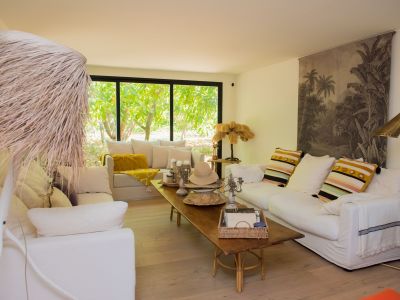 Amplia sala de estar con suelos de madera clara y sofás blancos. Grandes ventanales enmarcan vistas a un exuberante jardín tropical, realzados por una mesa de centro rústica y lámparas decorativas únicas.