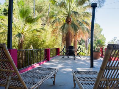 Amplia terraza exterior con dos tumbonas de bambú y un conjunto de mesa y sillas, enmarcada por un exuberante jardín tropical con palmeras y barandales fucsia. El suelo de hormigón recibe luz natural.