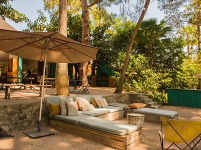Amplia zona de estar exterior con sofás de madera y cojines celestes, bajo una gran sombrilla. El suelo de grava se integra en un jardín tropical exuberante con palmeras e iluminación ambiental.