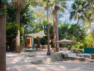 Amplio patio ajardinado tropical con varias zonas de asientos sombreadas sobre grava, incluyendo una gran mesa de comedor bajo un toldo con luces y un área lounge acolchada.
