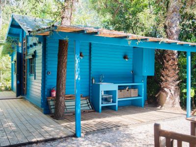 Cabaña de madera azul vibrante con cocina exterior integrada, fregadero y estantes abiertos, sobre una tarima de madera. Árboles tropicales crecen a través de su dosel natural, rodeada de caminos de grava.