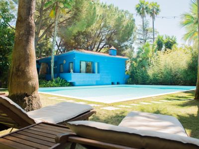 Casa azul vibrante con tejado de terracota, junto a una piscina de agua clara. Dos tumbonas de madera sobre el césped cuidado, enmarcadas por palmeras tropicales y vegetación frondosa bajo luces festivas.