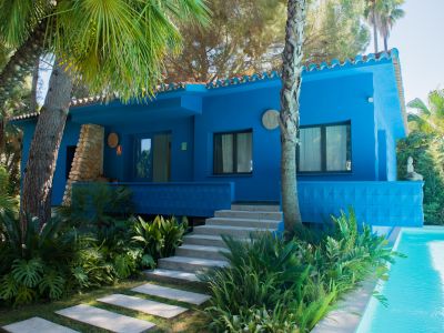 Casa azul vibrante con tejado tradicional, rodeada de exuberantes jardines tropicales y palmeras. Escaleras de hormigón conducen a una terraza elevada con vistas a una serena piscina.