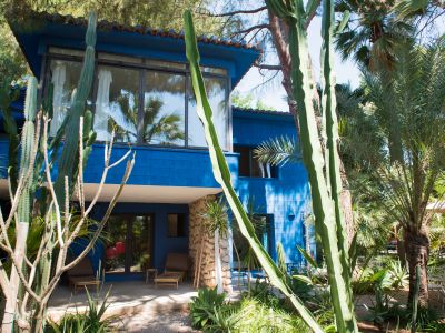 Casa de dos plantas en azul vibrante con patio sombreado en planta baja, tumbonas de bambú y pilares de piedra. El jardín tropical está enmarcado por cactus altos y palmeras.
