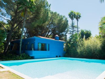 Casa moderna azul con tejado de terracota y una serena piscina con borde claro. Rodeada de exuberante vegetación tropical, incluyendo pinos y palmeras, bajo un cielo despejado.