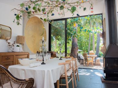 Comedor ecléctico con mesa formal y candelabros de cristal, frente a puertas de vidrio con vista a jardín tropical. Una instalación de ramas en el techo con luces y una chimenea moderna completan el espacio.