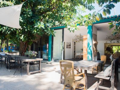 Comedor exterior con mesas de madera y sillas variadas bajo una vela de sombra blanca, enmarcado por exuberante vegetación tropical y conectado al interior por puertas de cristal.