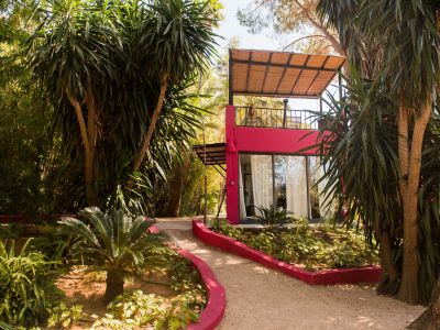 Edificio moderno color fucsia con amplias puertas de cristal, dosel de material natural tejido y balcón superior con asientos de ratán. Un sendero de grava serpentea entre la exuberante vegetación tropical.