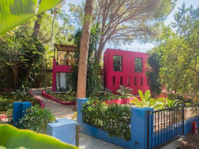 Edificio moderno de color fucsia vibrante con ventanas variadas, enclavado en un denso jardín tropical con árboles altos y senderos serpenteantes. Un muro bajo azul con arbustos en flor enmarca el primer plano.