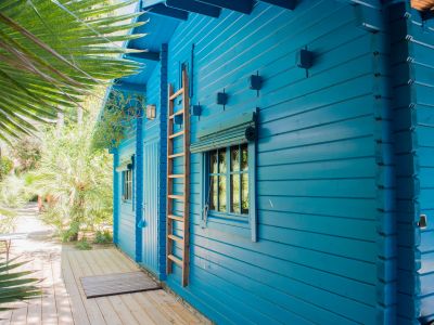 Exterior de cabaña de madera azul con escalera decorativa y ventana con persiana de bambú. Una terraza de madera clara se integra en un exuberante jardín tropical.