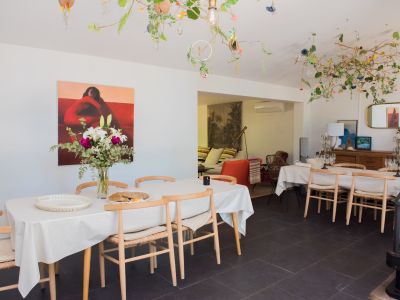 Luminoso comedor con mesa larga, sillas de madera clara y arreglo floral fresco. El techo exhibe una instalación natural única con ramas, flores y luces sutiles, sobre un suelo de baldosas oscuras.