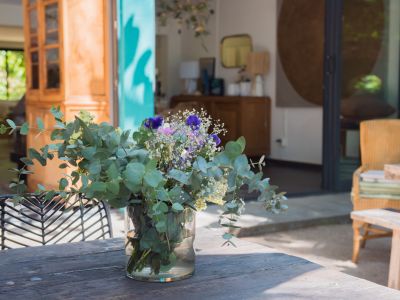 Mesa exterior rústica de madera con un fresco arreglo floral de eucalipto, enmarcada por una puerta turquesa abierta que conecta con un interior luminoso y un jardín exuberante.