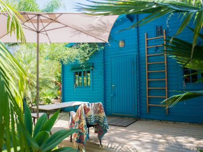 Terraza de madera con sombrilla y mobiliario exterior, destacando una vibrante cabaña azul con escalera de madera, inmersa en un exuberante jardín tropical.