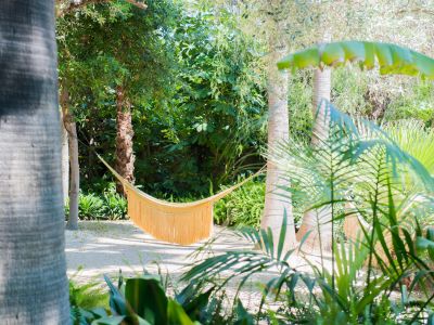 Una hamaca amarilla con flecos ofrece un rincón sereno para el descanso, entre dos palmeras en un jardín tropical soleado con sendero de grava clara.