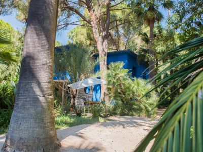 Vista exterior de una casa azul vibrante con detalles de piedra, rodeada por un exuberante jardín tropical. Un área de asientos al aire libre con sillas blancas y sombrilla se ve en un sendero de grava.